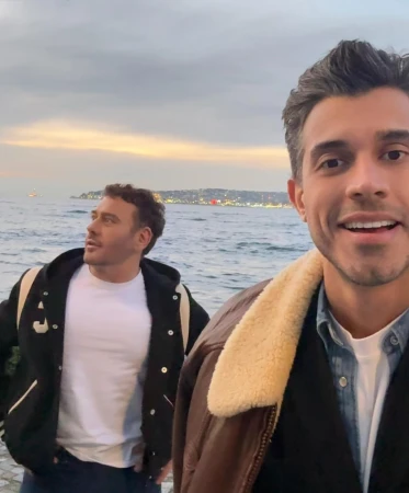Two men outdoors by the water at sunset, one smiling in the foreground wearing a brown jacket with a shearling collar.