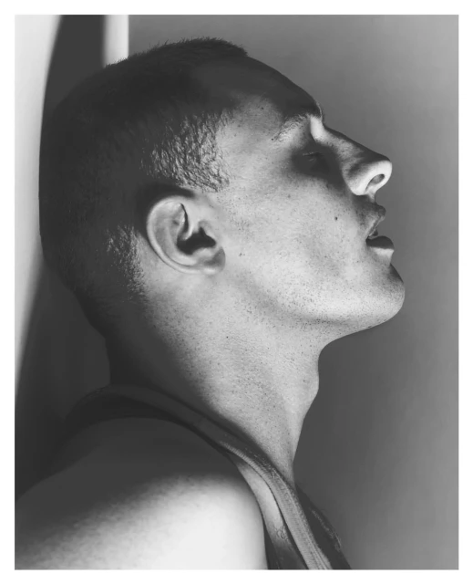 Black and white portrait of a young adult male with short hair, resting against a wall in an indoor setting with neutral lighting.