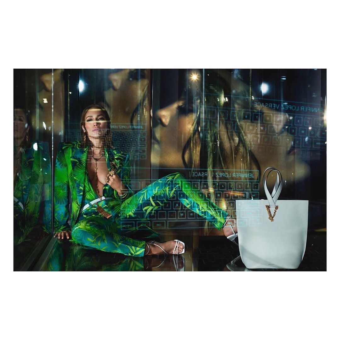 Jennifer Lopez poses in a green patterned suit next to a white Versace handbag against a reflective tech background with a face overlay.