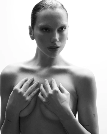 Portrait of a woman with wet-look hair and tattoos on her hands posing against a bright white background.