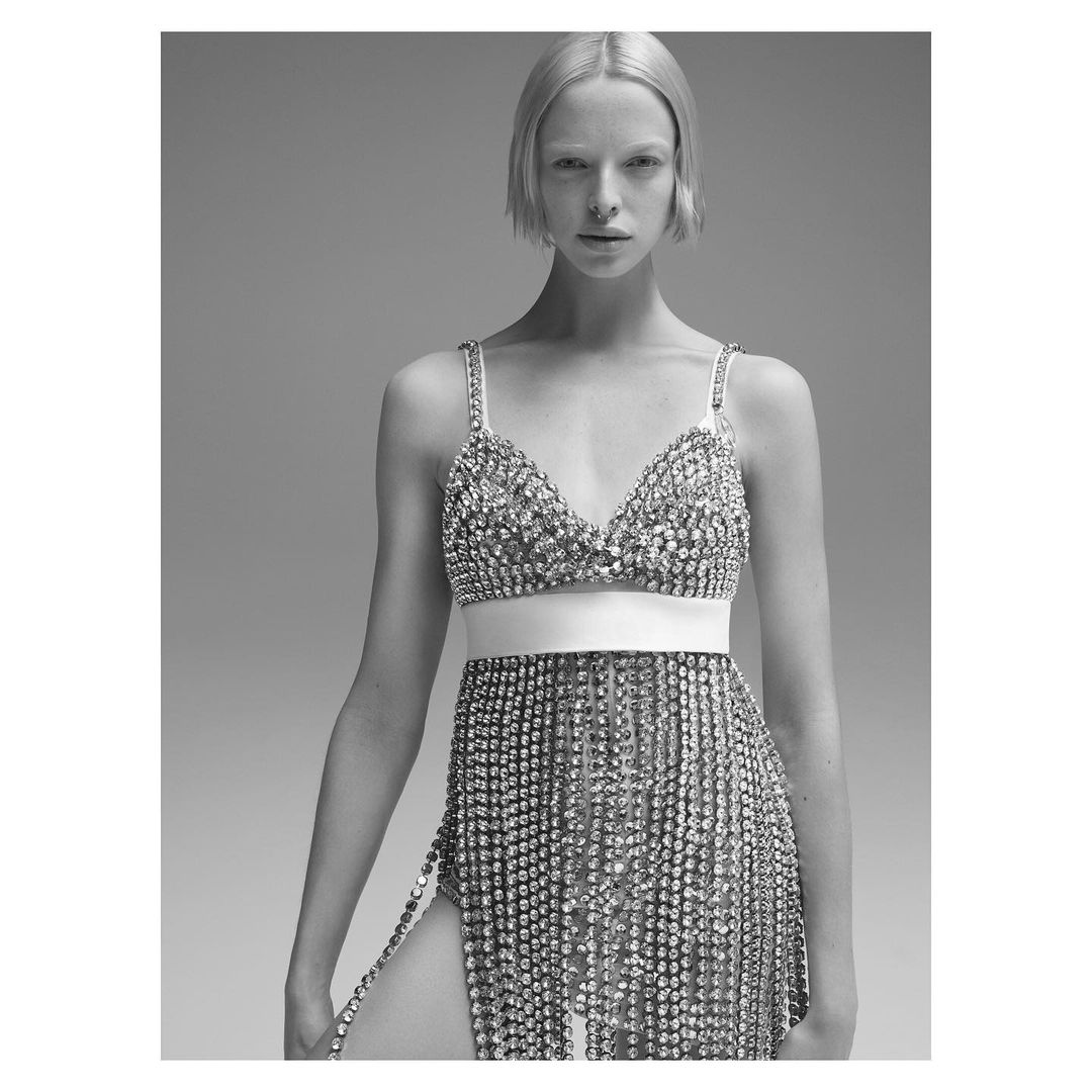 Black and white portrait of a woman in a rhinestone bra top and matching skirt, posing against a grey backdrop with a neutral expression.