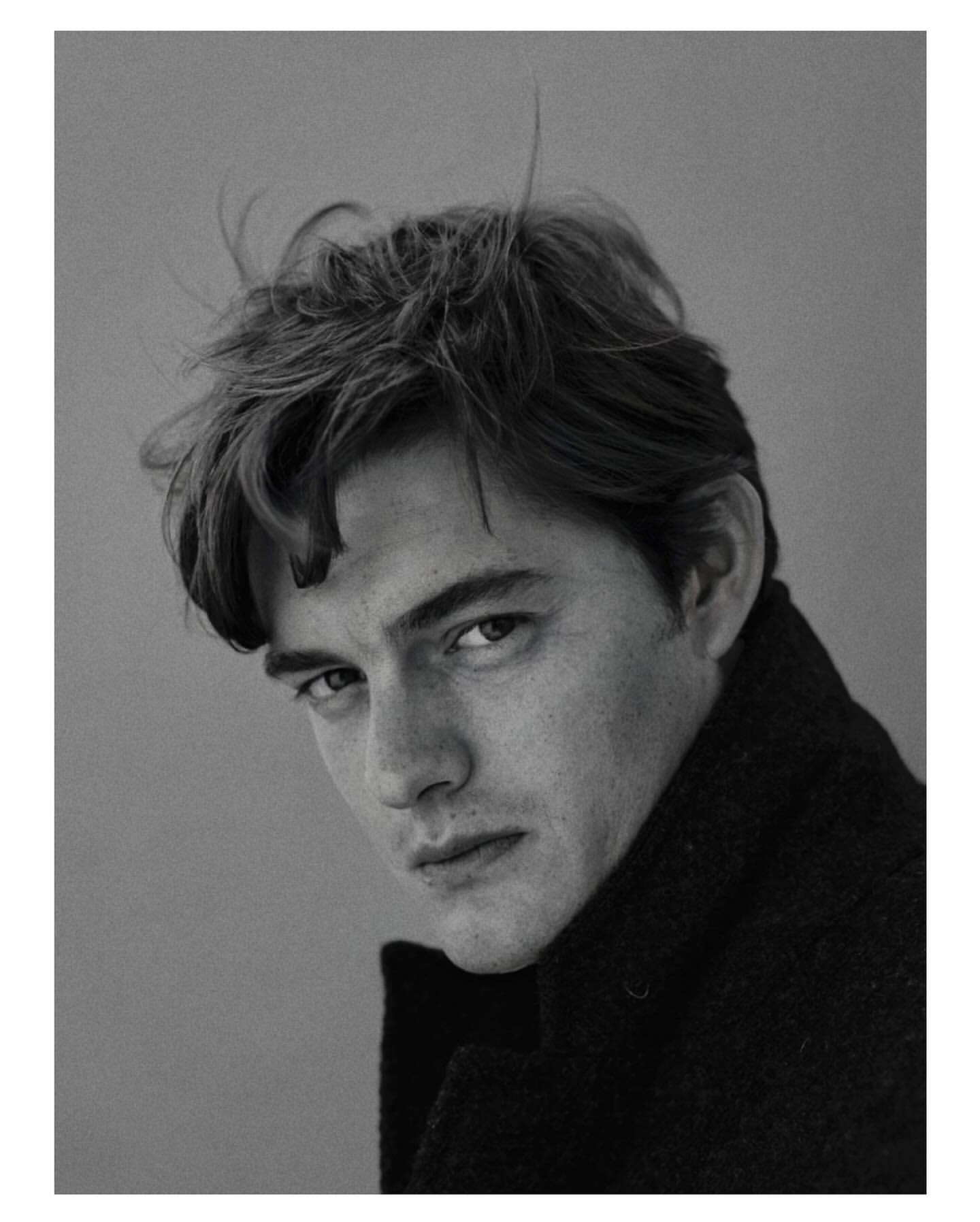 Monochromatic portrait of a dark-haired adult male gazing toward the camera while wearing a black textured coat against a grey backdrop.