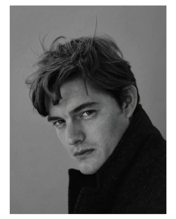 Monochromatic portrait of a dark-haired adult male gazing toward the camera while wearing a black textured coat against a grey backdrop.