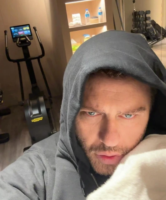 Man in a grey hooded sweatshirt in a fitness room with exercise equipment, water bottles and towels. Posing indoors in a home gym.