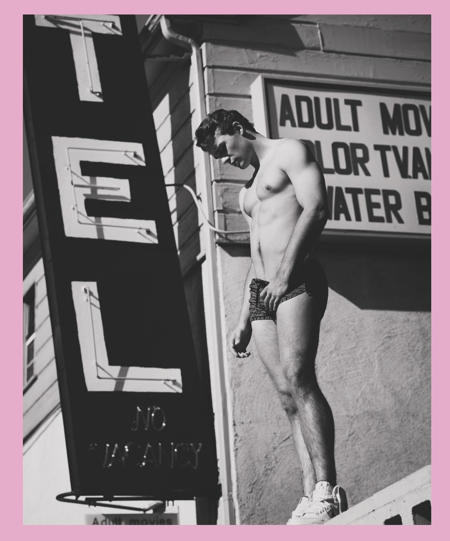 Athletic man poses shirtless outside a motel with vintage signage in a black and white outdoor shot showing shadow and sun.