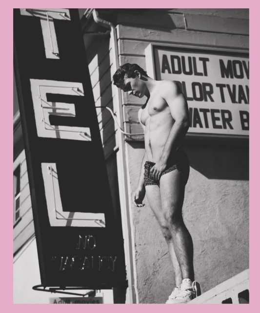 Athletic man poses shirtless outside a motel with vintage signage in a black and white outdoor shot showing shadow and sun.