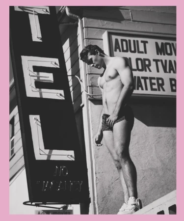 Athletic man poses shirtless outside a motel with vintage signage in a black and white outdoor shot showing shadow and sun.
