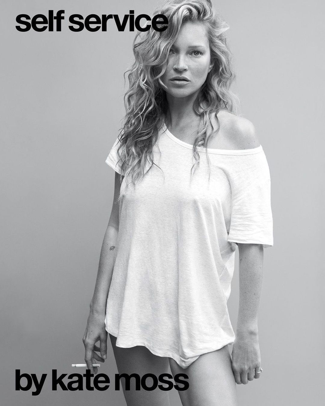 Kate Moss poses in a black-and-white portrait, wearing a white t-shirt, with curly blonde hair and a cigarette in her left hand.