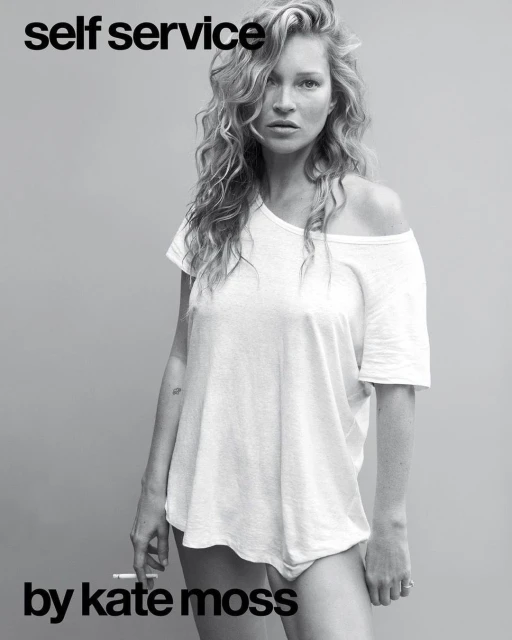 Kate Moss poses in a black-and-white portrait, wearing a white t-shirt, with curly blonde hair and a cigarette in her left hand.