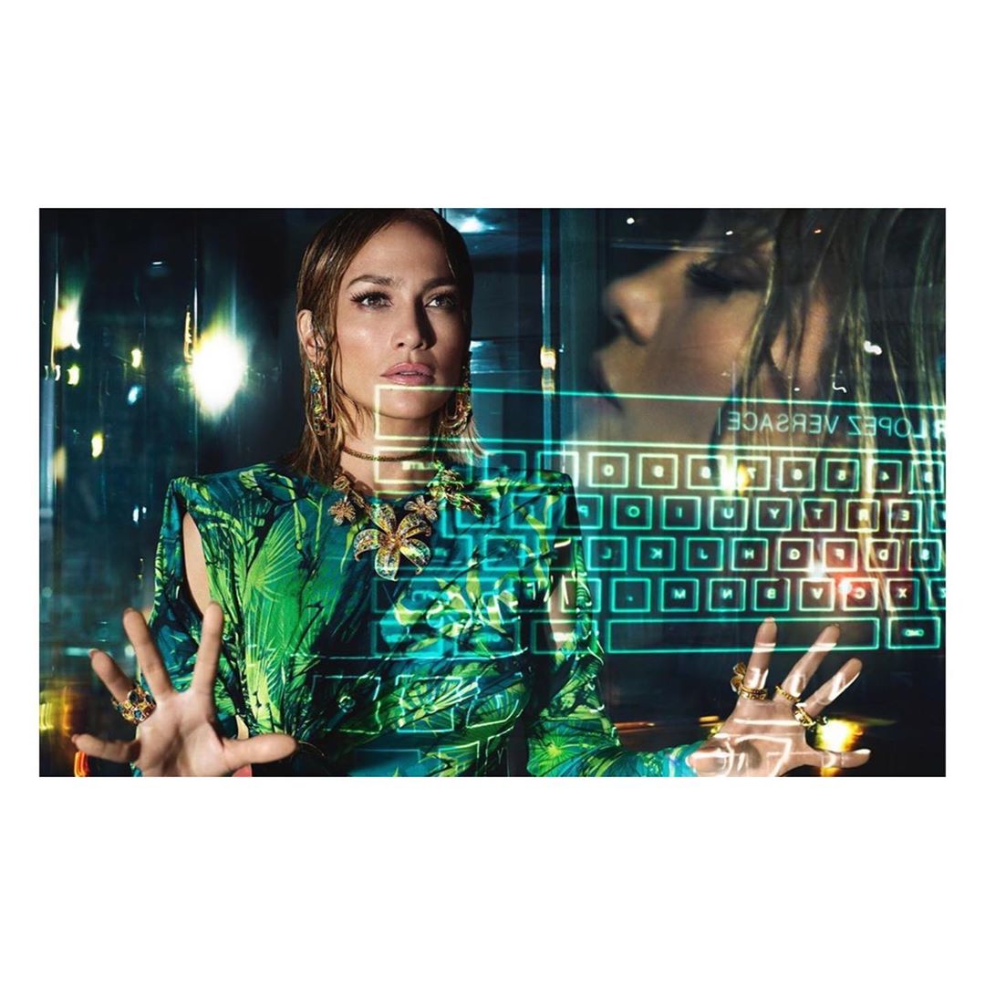 Jennifer Lopez poses in a green Versace dress and gold jewelry in an indoor setting with a futuristic, digital keyboard overlay.