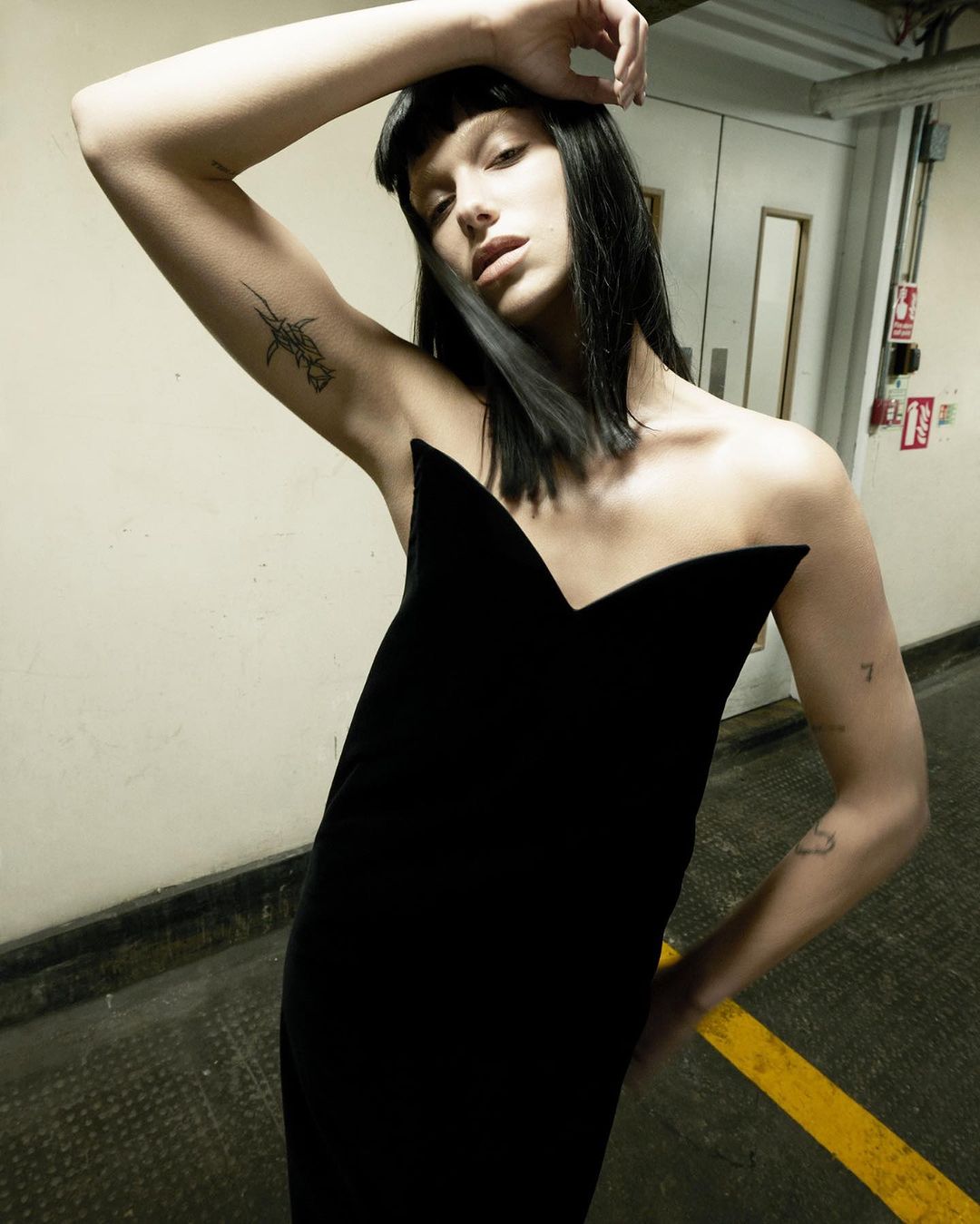 A woman with dark hair wearing a black velvet dress and posing indoors against a wall in a high-fashion editorial style.