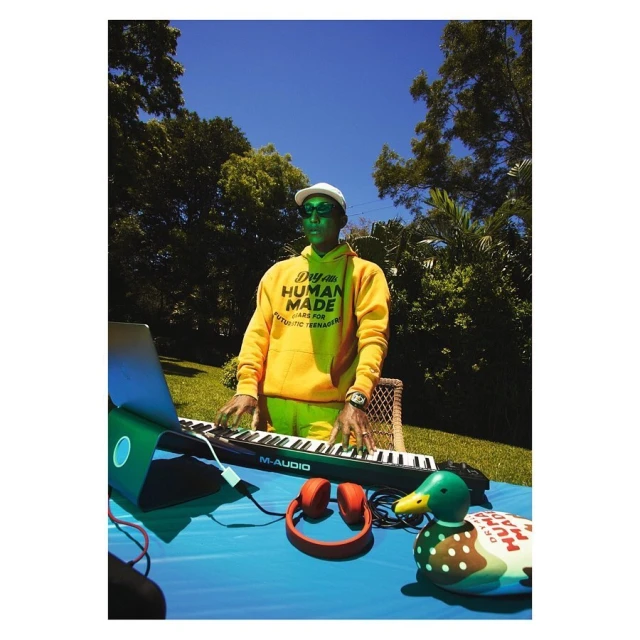 Man in yellow hoodie plays keyboard outdoors on a table with laptop, headphones, and duck figure under a bright blue sky.