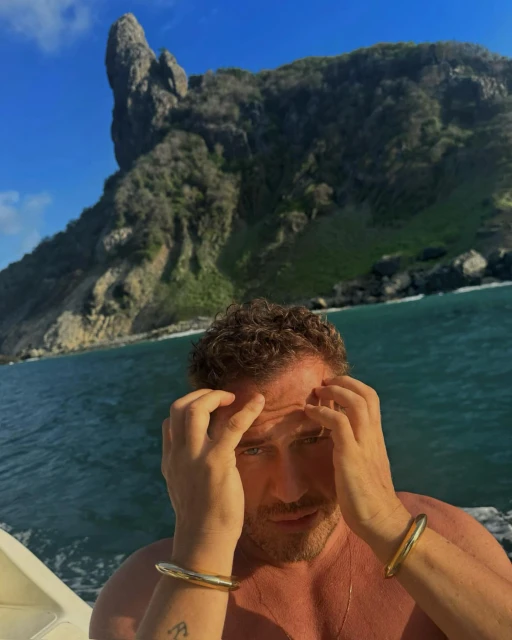 Adult man with curly brown hair poses in front of a tropical island and turquoise sea, wearing gold cuff bracelets and necklace.