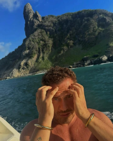 Adult man with curly brown hair poses in front of a tropical island and turquoise sea, wearing gold cuff bracelets and necklace.