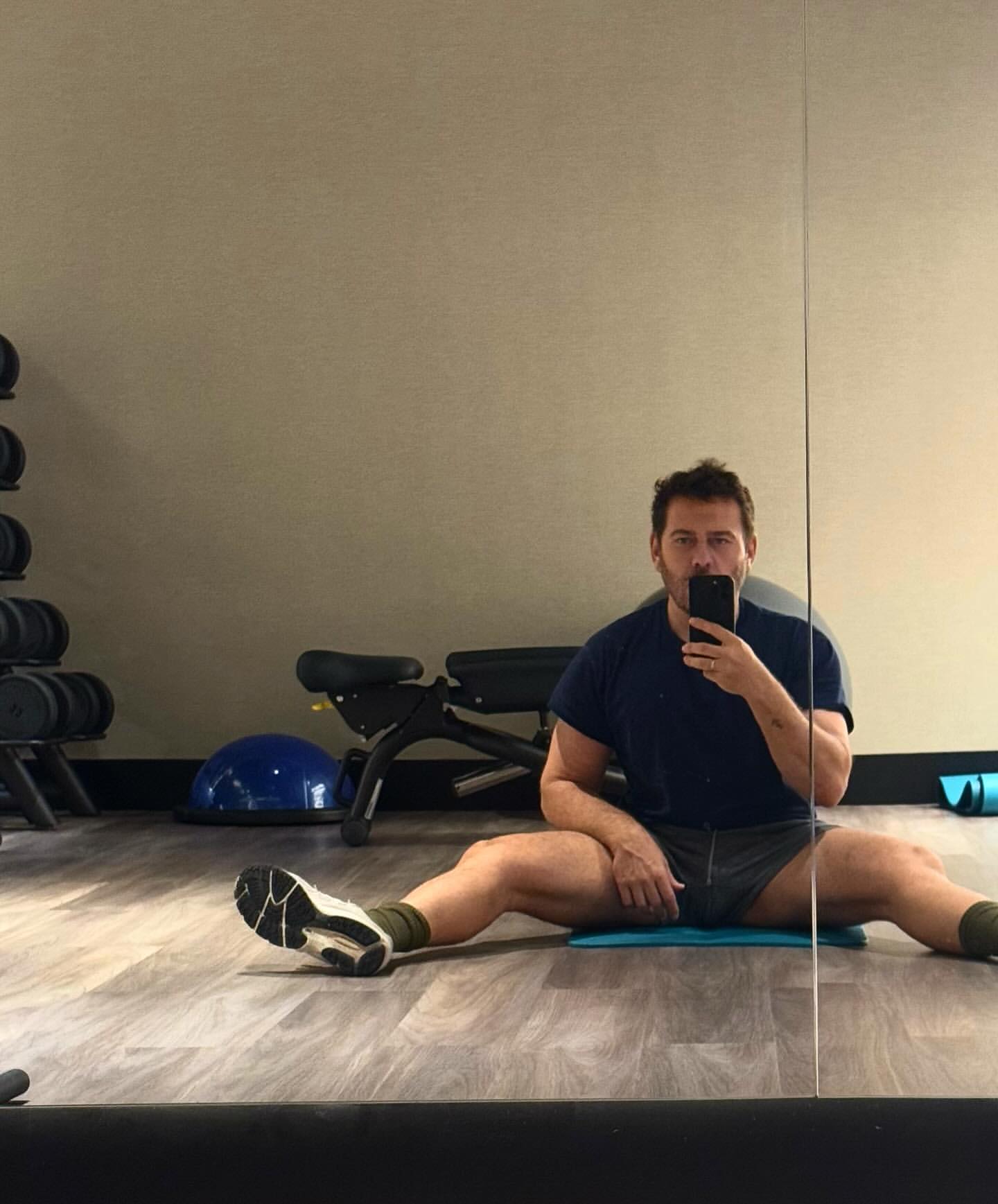 Man in sportswear does splits, using his phone at an indoor gym with weights, exercise ball, rubber mats, and wooden floors.
