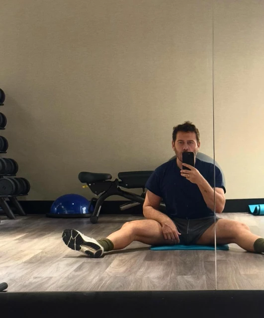 Man in sportswear does splits, using his phone at an indoor gym with weights, exercise ball, rubber mats, and wooden floors.