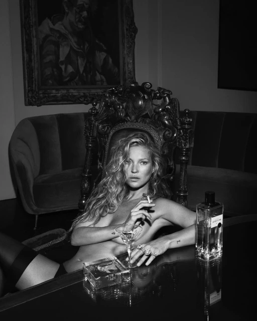 Black and white photo of a woman sitting in an ornate chair, holding a cigarette with a glass and a perfume bottle on the table.
