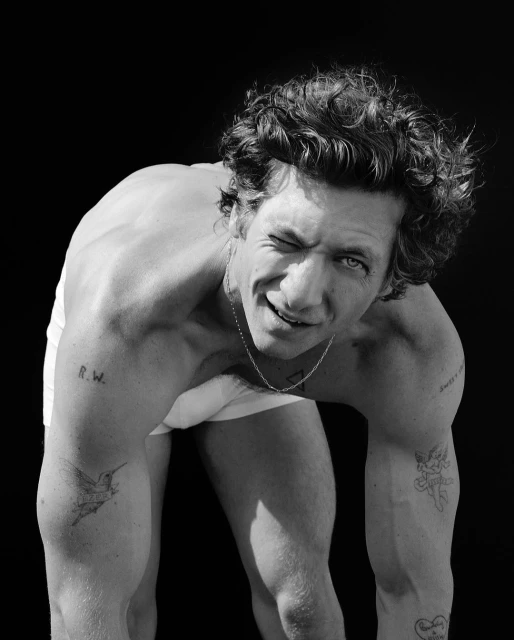 Black and white portrait of a shirtless, light-skinned adult male winking at the camera with tattoos on his arms against black backdrop.
