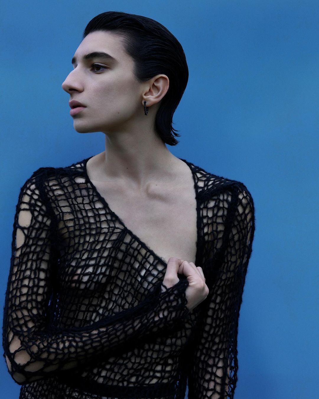 Close-up studio shot of a slim adult with short dark hair, wearing a black open-weave sweater with a blue backdrop, gazing to the side.