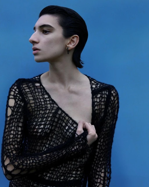 Close-up studio shot of a slim adult with short dark hair, wearing a black open-weave sweater with a blue backdrop, gazing to the side.