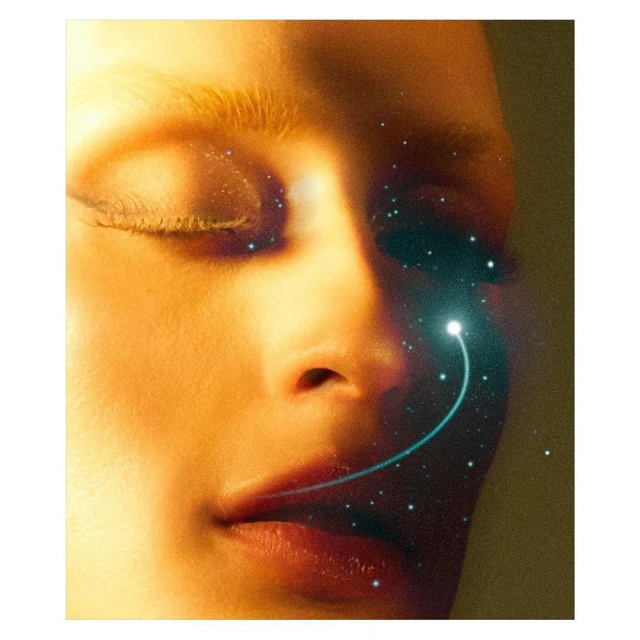 Close-up beauty shot of an adult female face, lit in golden tones with a sparkling, glowing light effect near her nose and eye area.