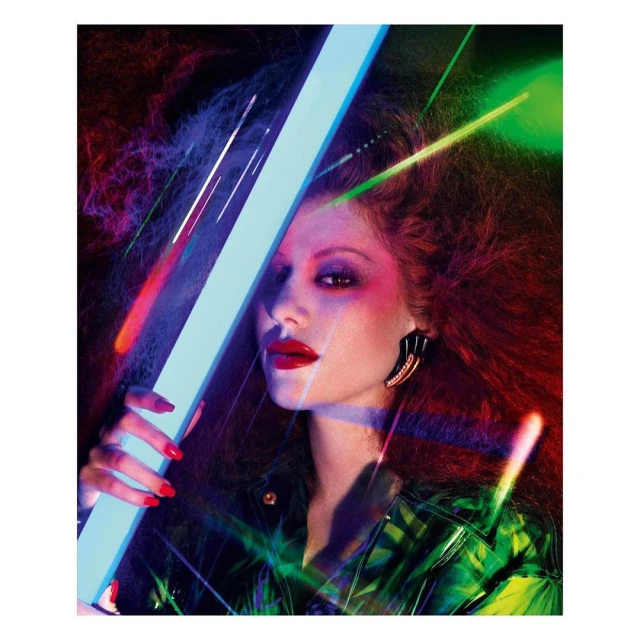 Close-up of a stylish adult female with red hair, red lipstick, and bold earrings holding a glowing blue light tube in a dark studio.