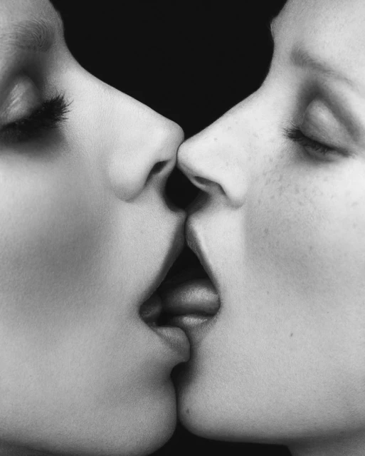 Close-up grayscale image of two faces nearly touching, emphasizing the lips and noses in a symmetrical, intimate composition against a black backdrop.