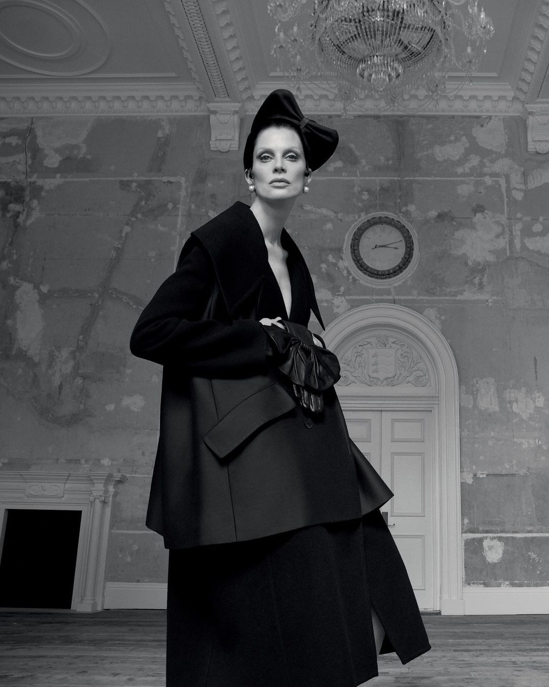 Elegant woman poses in black couture dress and bow hat in a derelict room with white door and chandelier in black and white photography.