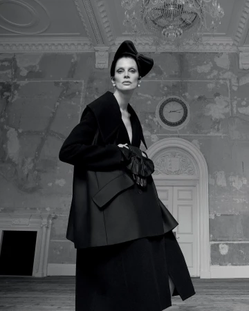 Elegant woman poses in black couture dress and bow hat in a derelict room with white door and chandelier in black and white photography.