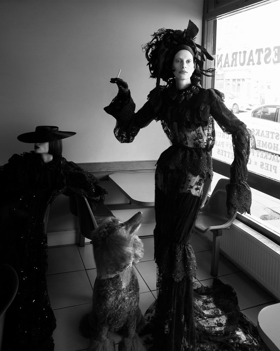 Black and white image showcasing two figures in elaborate dark dresses, one smoking, with a dog-like sculpture in an indoor restaurant setting.