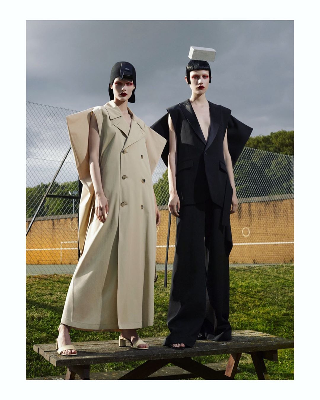 Two models pose in avant-garde fashion outdoors; woman in tan double-breasted coat, another in black suit and hat; both with head accessories.