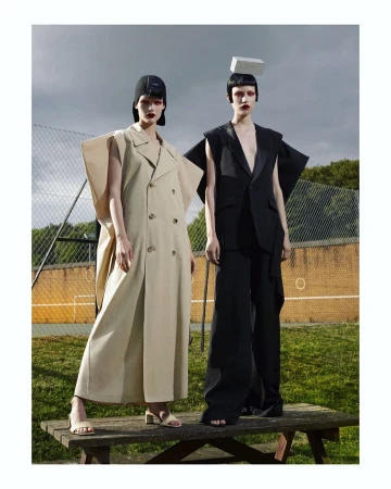 Two models pose in avant-garde fashion outdoors; woman in tan double-breasted coat, another in black suit and hat; both with head accessories.