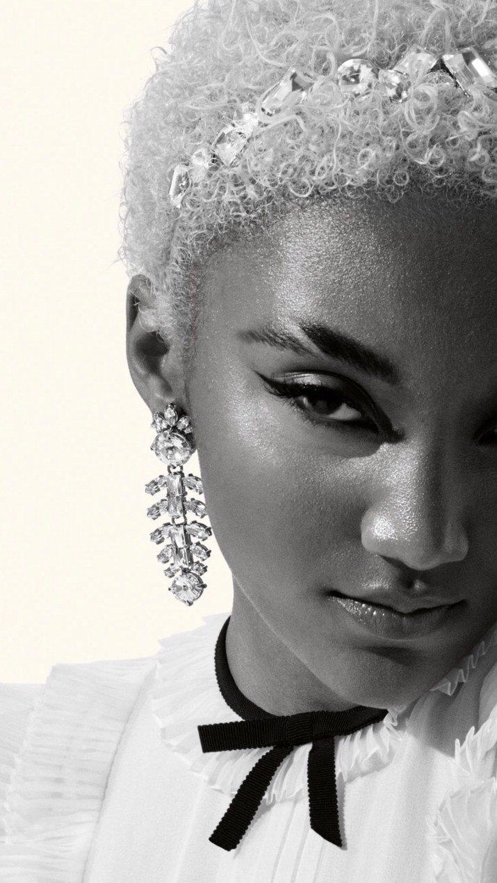 Close-up portrait of a Black woman with platinum blonde hair and statement earrings, wearing a black ribbon choker and white collared shirt.