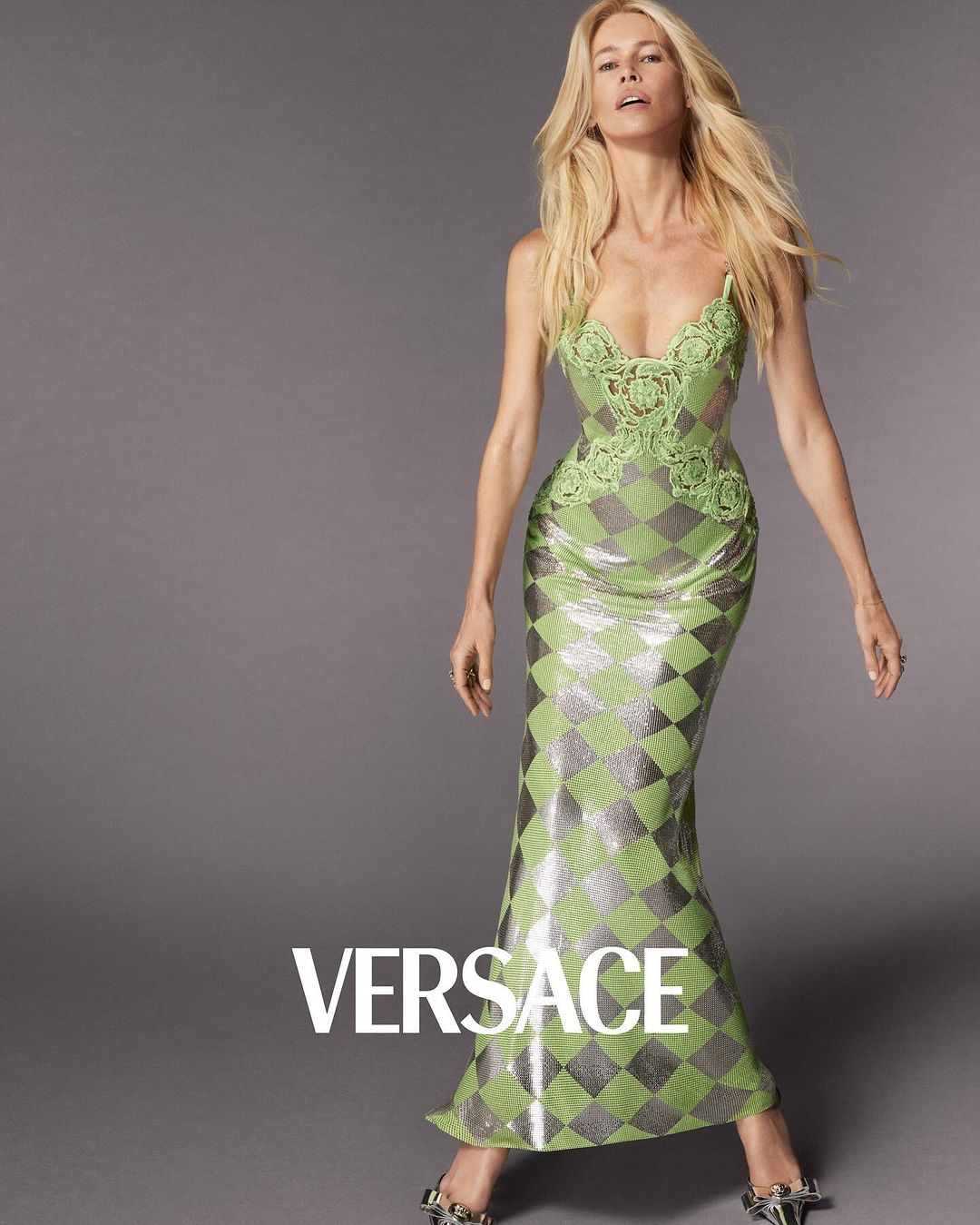 Fashion shot of a blonde woman in a green sequined gown with lace details, posing against a grey background. Versace logo visible.