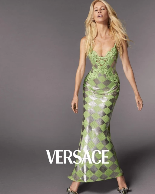 Fashion shot of a blonde woman in a green sequined gown with lace details, posing against a grey background. Versace logo visible.