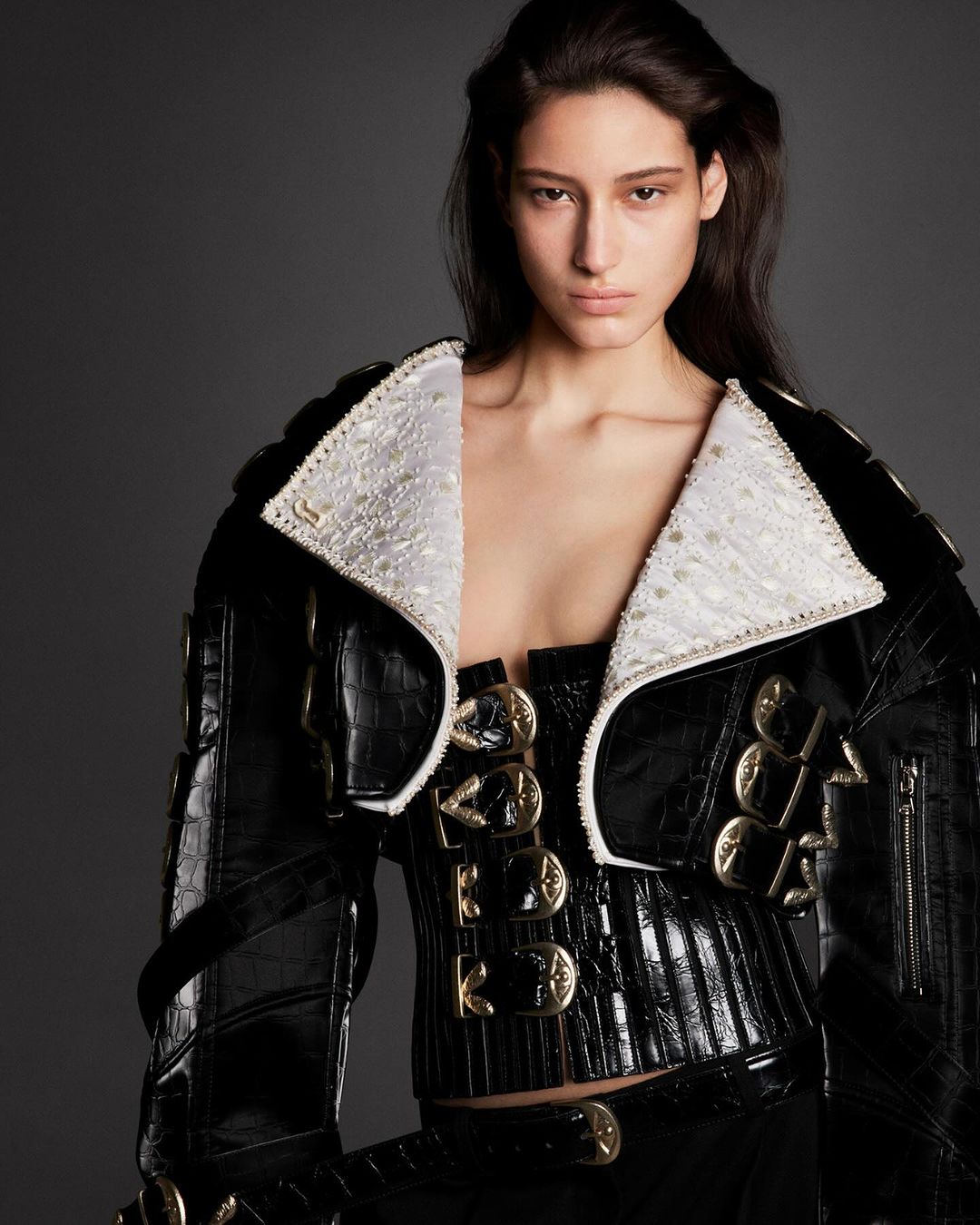 Portrait of a fashion model wearing a black leather jacket with a white collar, a corset top, and a matching skirt in a studio setting.