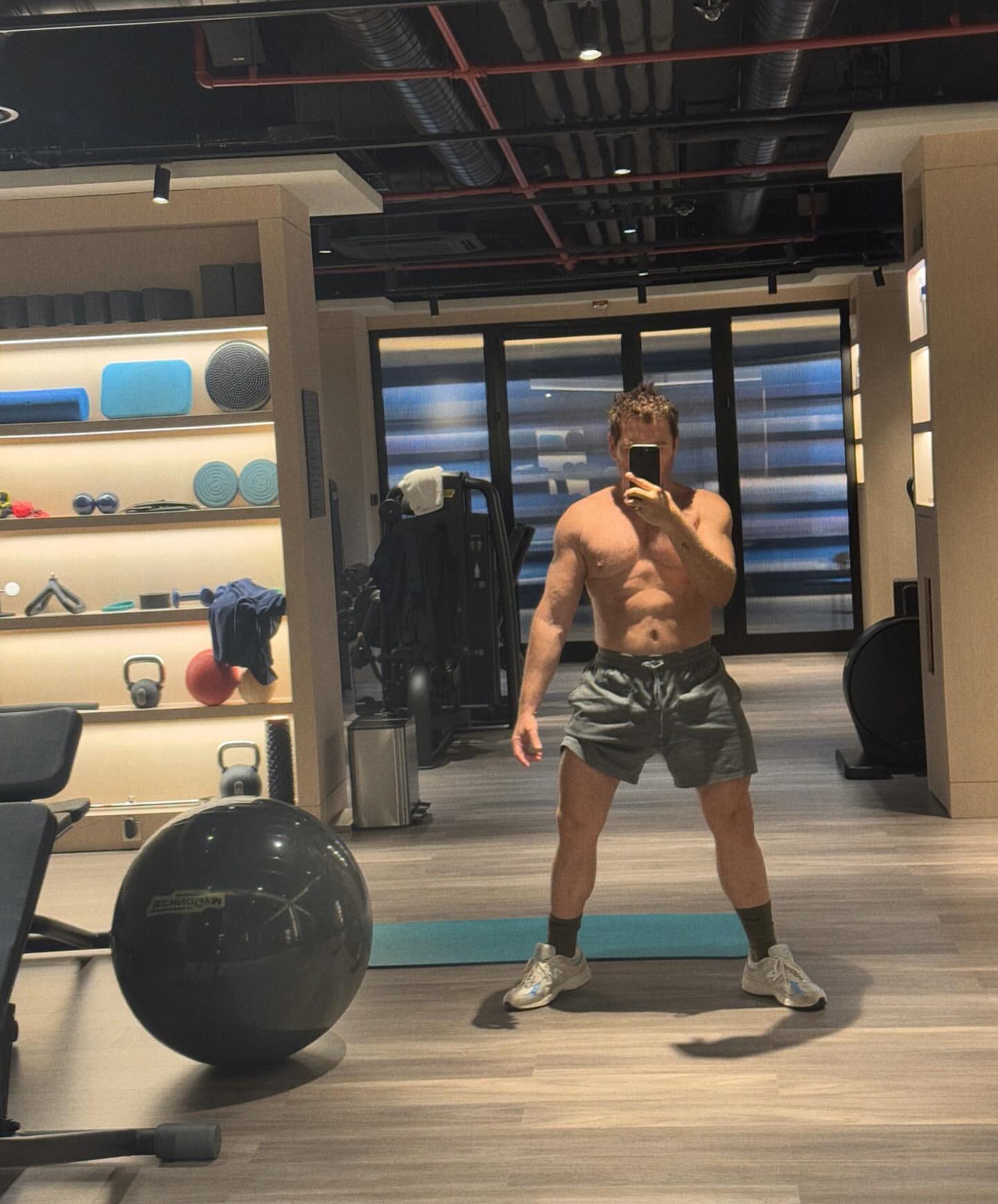 Adult male in a fitness center gym takes a selfie, wearing athletic shorts and shoes with exercise equipment and a mirror background.