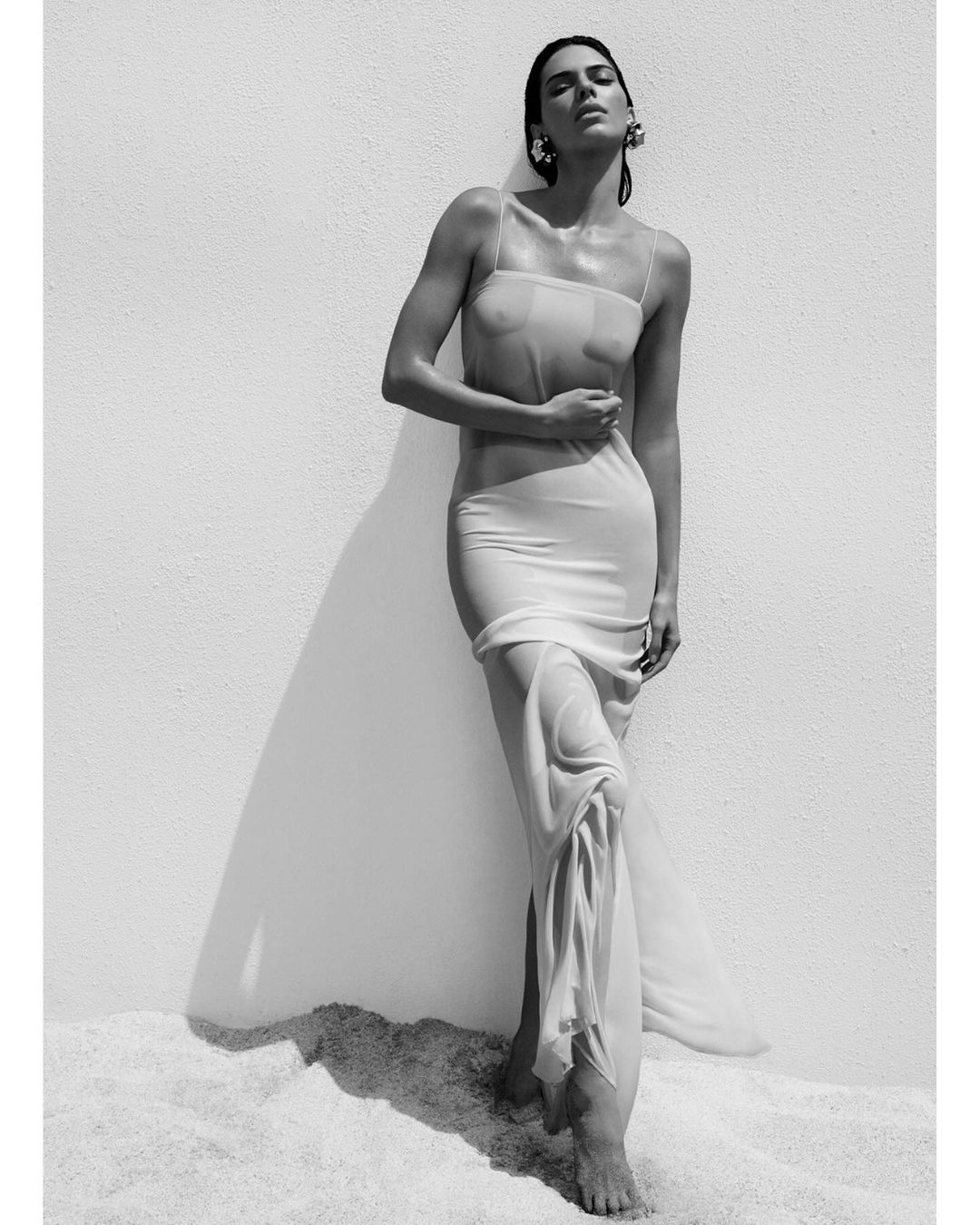 Black and white portrait of a woman wearing a sheer slip dress standing against a light wall on sand