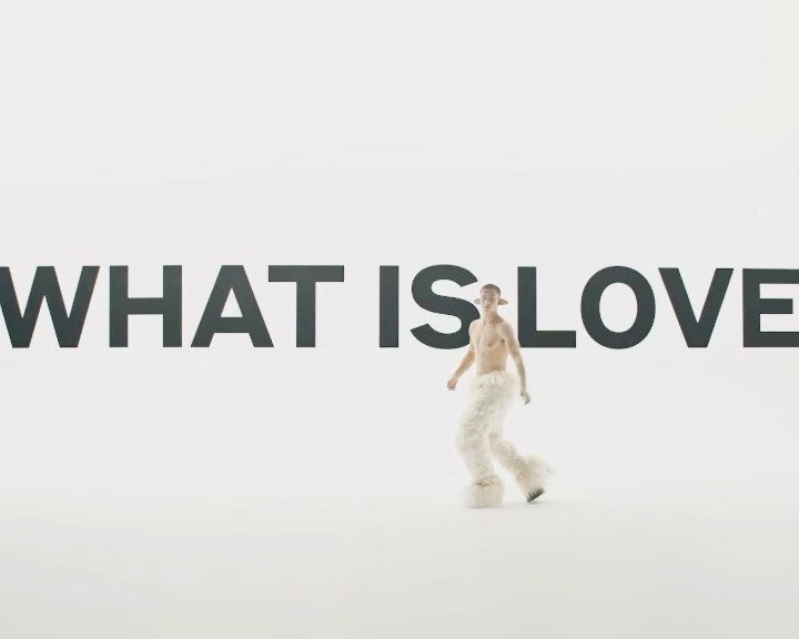Person in fur pants and ears posing in front of the text graphic 'What is Love' in a brightly lit white photo studio backdrop.