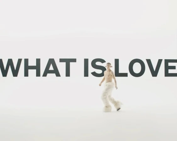 Person in fur pants and ears posing in front of the text graphic 'What is Love' in a brightly lit white photo studio backdrop.