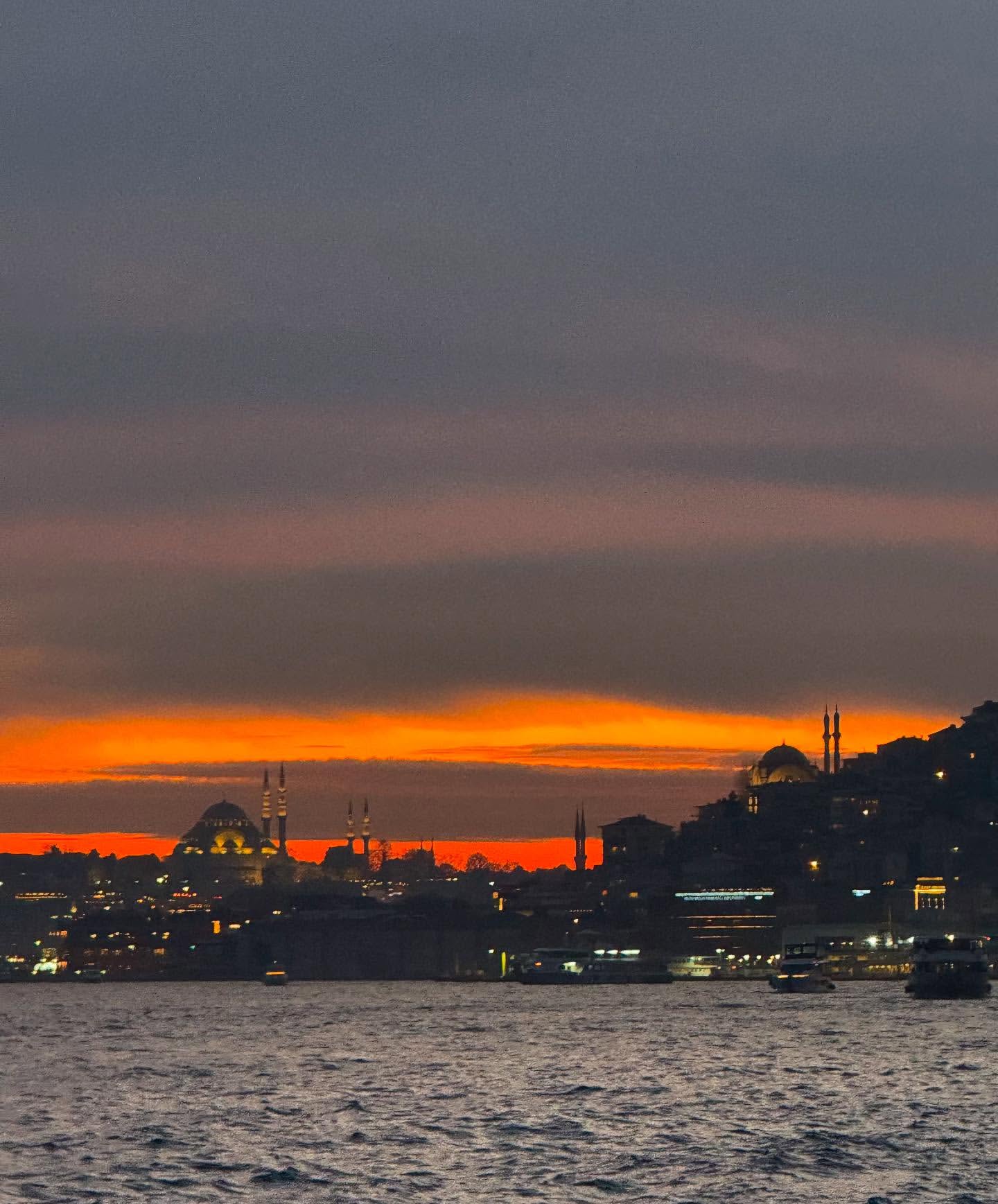 City skyline at dusk with silhouettes of mosques and buildings along the water, lit by the colorful glow of the setting sun.