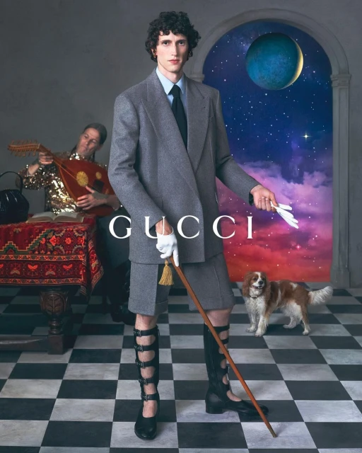 An androgynous fashion model poses in a tailored grey suit with knee-high buckle boots, and a cane next to a mandolin player.