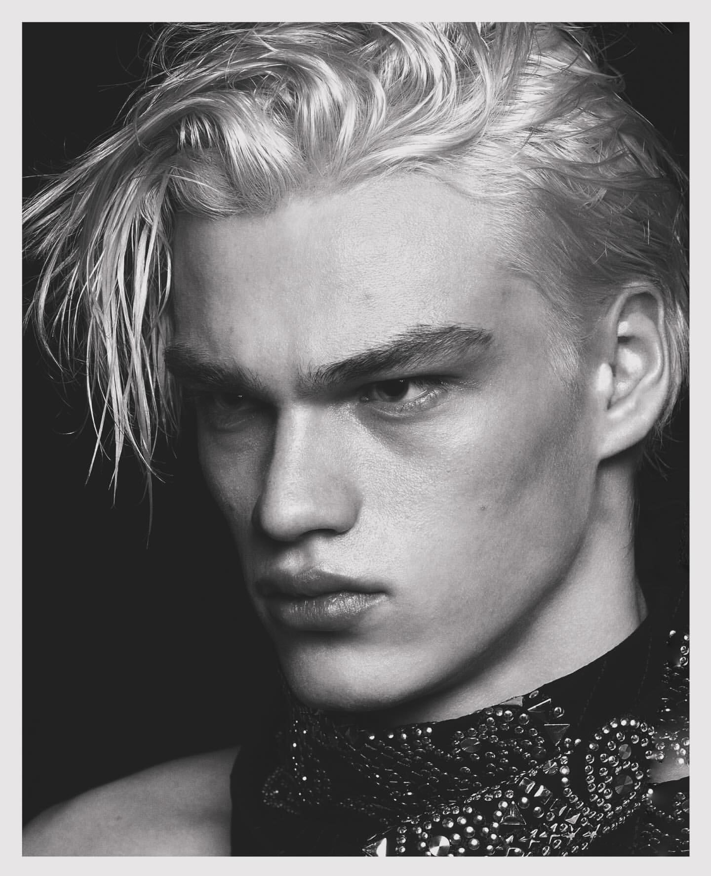 Black and white portrait of a young adult male model with dyed blond hair, wearing a studded, textured neck piece, against a dark background.