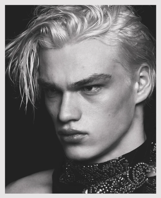 Black and white portrait of a young adult male model with dyed blond hair, wearing a studded, textured neck piece, against a dark background.
