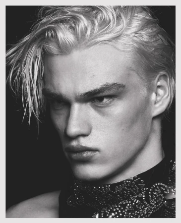 Black and white portrait of a young adult male model with dyed blond hair, wearing a studded, textured neck piece, against a dark background.