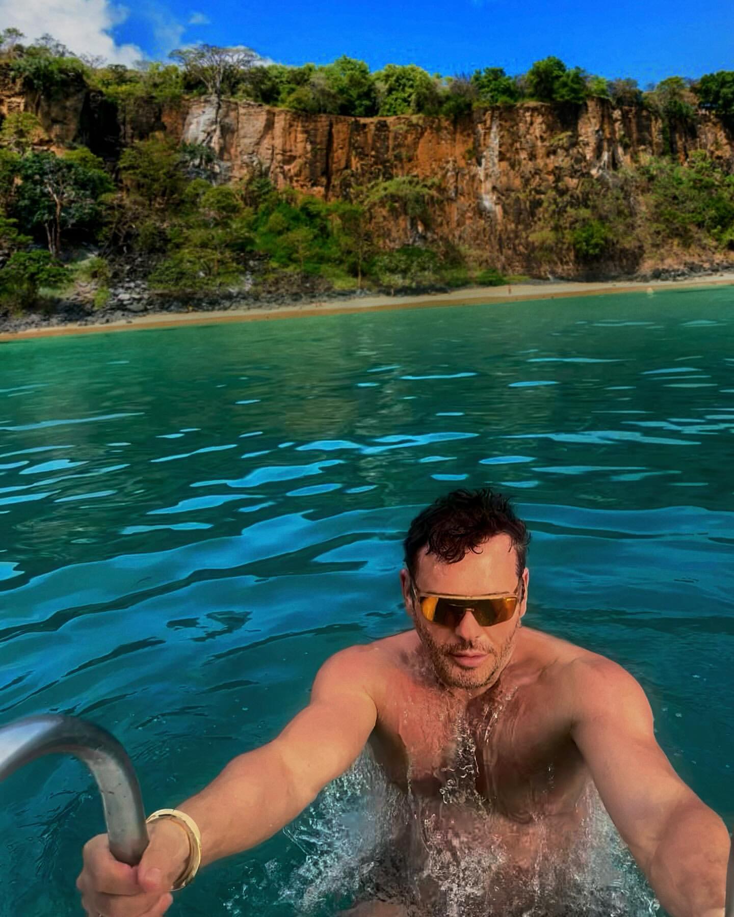 Man wearing sunglasses swimming in bright turquoise ocean water with a rocky cliff in the background
