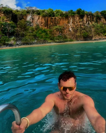 Man wearing sunglasses swimming in bright turquoise ocean water with a rocky cliff in the background