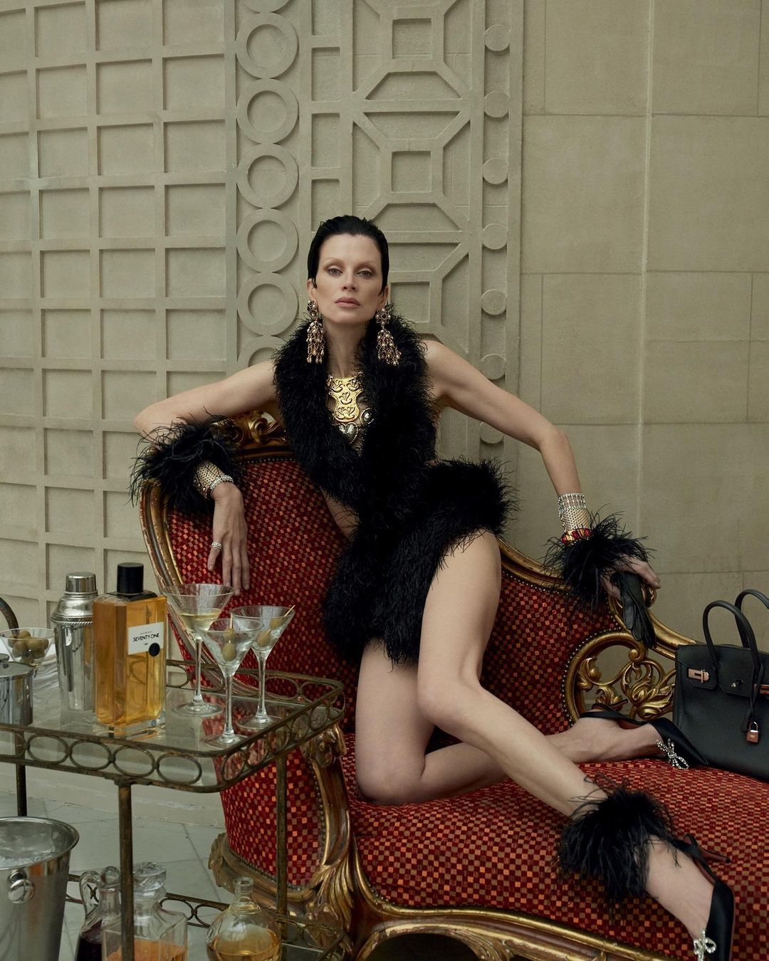 Woman posing in feather dress and jewelry on chaise lounge with bar cart, drinks, and purse against a decorative architectural wall.