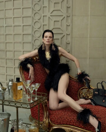 Woman posing in feather dress and jewelry on chaise lounge with bar cart, drinks, and purse against a decorative architectural wall.