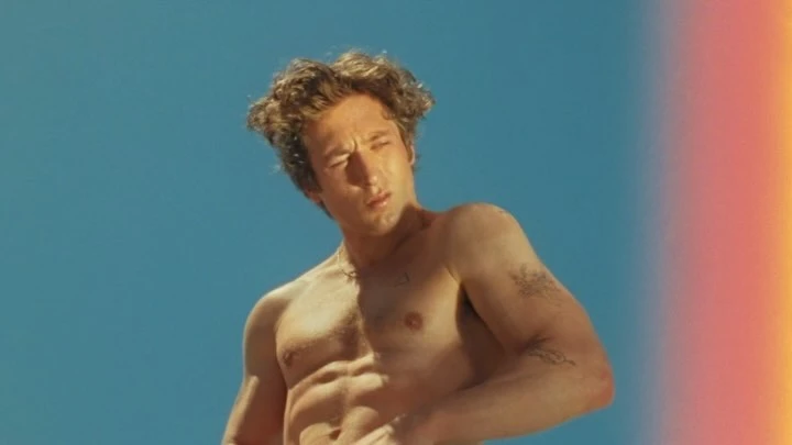 Shirtless man with curly brown hair poses against a blue sky, showing his physique with a relaxed expression in outdoor environment.